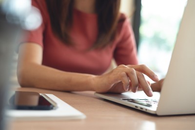A woman with brown hair in a coral coloured t-shirt, typing on a laptop keyboard